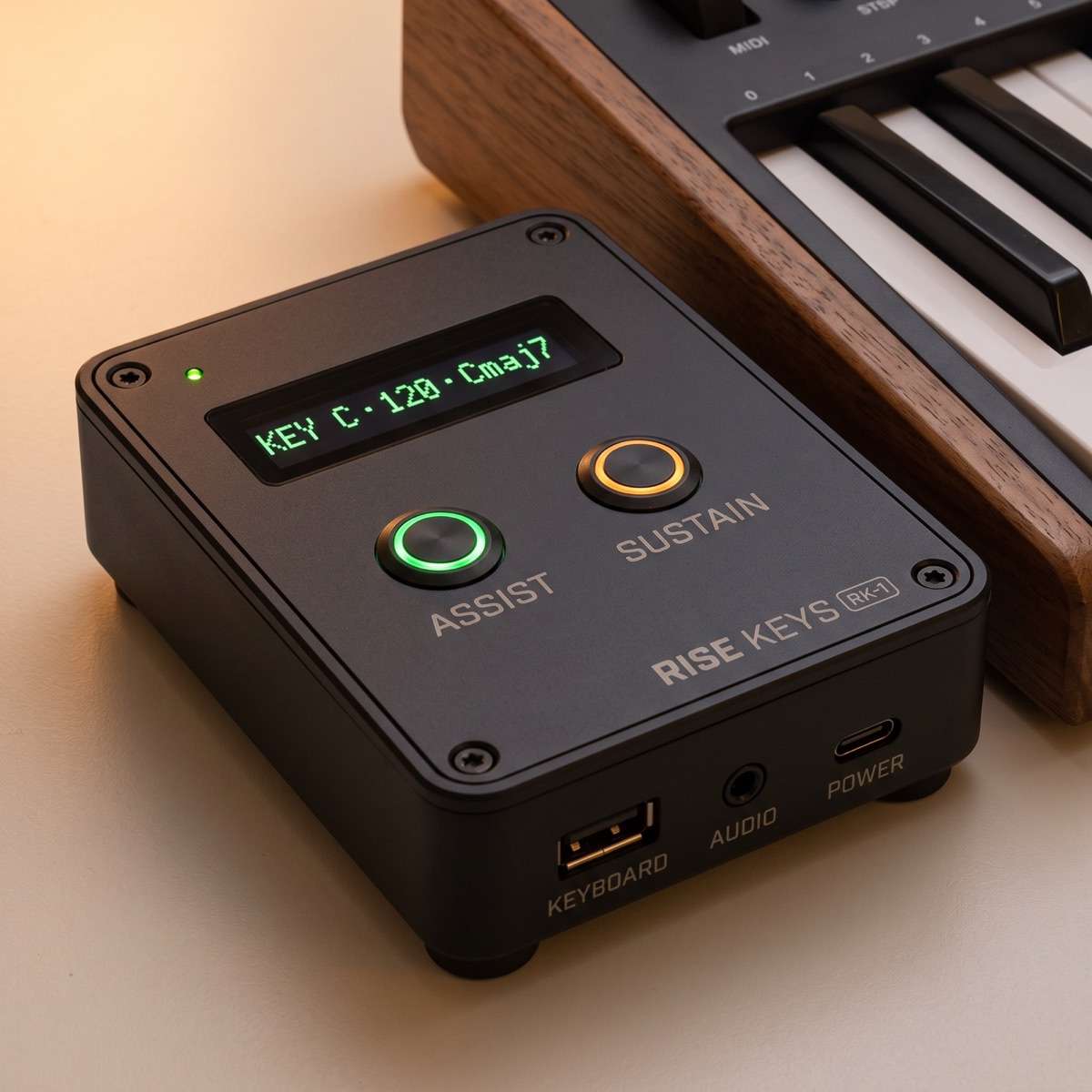 Rise Keys RK-1: a matte charcoal box with a
    green OLED display, glowing ASSIST and SUSTAIN buttons, beside a
    MIDI keyboard
